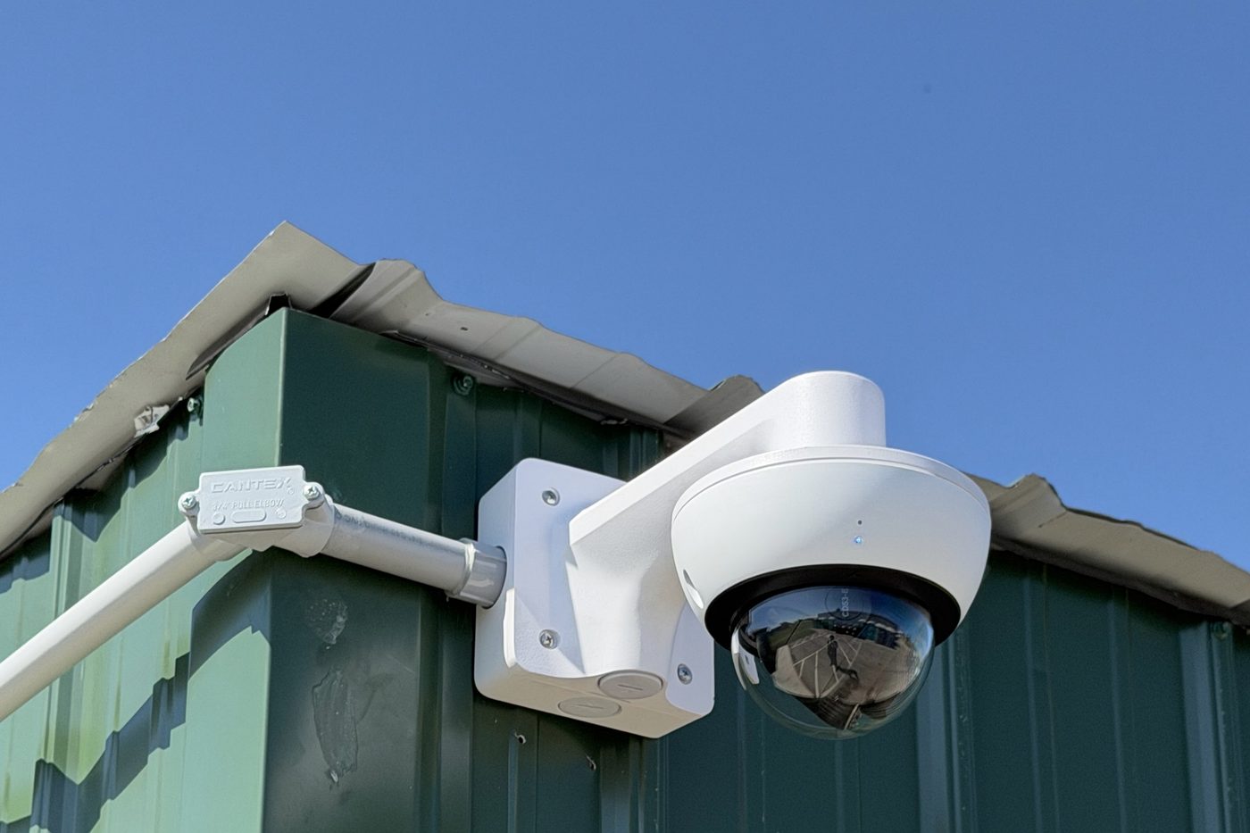 Verkada outdoor dome camera mounted at the roofline of a metal building, with conduit run into a sealed back box